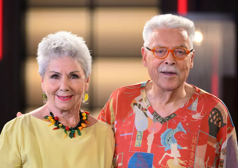 Martina und Moritz lächeln in die Kamera vor unscharfem Hintergrund. Sie tragen ein gelbes Oberteil und eine bunte Kette bzw. ein buntes Oberteil und eine orangene Brille.