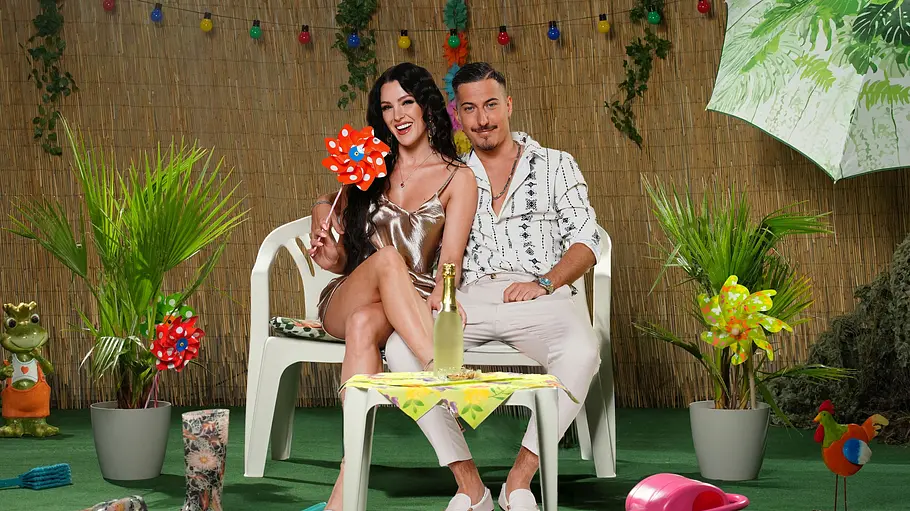 Marvin Kleinen und Jennifer Degenhart Marvin Kleinen und Jennifer Degenhart sitzen eng beieinander zwischen Topfpalmen und bunter Gartendeko vor einer Schilfmatte.
