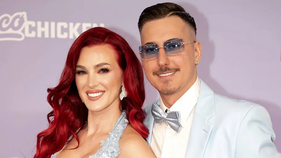 Marvin und Jenny stehen vor einem lila Hintergrund mit Schriftzug. Sie hat rote Haare, er trägt einen hellblauen Anzug und eine Sonnenbrille.