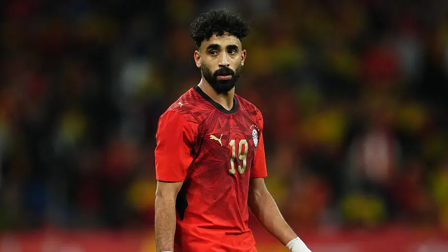 Ein Fußballspieler der ägyptischen Nationalmannschaft im roten Trikot mit der Rückennummer 19 steht während eines Spiels auf dem Rasen. Er blickt aufmerksam zur Seite, trägt Bandagen an beiden Handgelenken und vor unscharfem Stadionhintergrund.