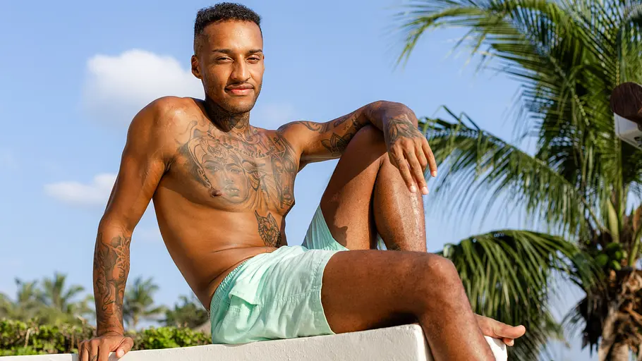 Marwin sitzt oberkörperfrei und mit einer hellblauen Shorts auf einem Stein und lächelt in die Kamera. Im Hintergrund sieht man blauen Himmel und Palmen.