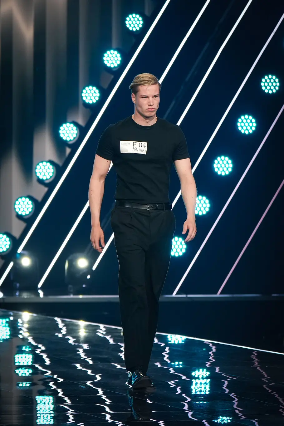 Ein männlicher GNTM-Kandidat läuft in schwarzem T-Shirt und schwarzer Hose selbstbewusst über den beleuchteten Laufsteg beim Entscheidungswalk.