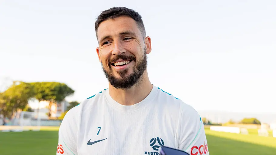 Ein australischer Nationalspieler im weißen Trainingsshirt steht lächelnd auf einem Fußballplatz und gibt Journalistinnen und Journalisten nach einer Trainingseinheit ein Interview. Im Vordergrund sind Mikrofone und Smartphones zu sehen.