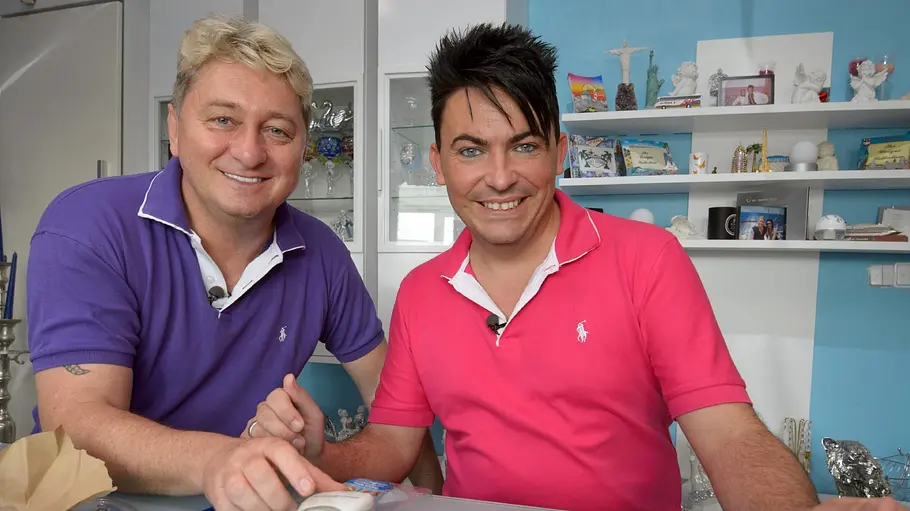 Hubert und Matthias Zwei Männer sitzen nebeneinander an einem Tisch und lächeln in die Kamera; vor ihnen liegen verschiedene kleine Produkte und Verpackungen, im Hintergrund sind Regale mit Dekoartikeln zu sehen.