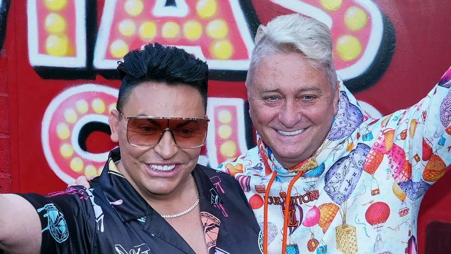 Matthias und Hubert Zwei Männer posieren lachend mit erhobenen Armen vor einer roten Wand mit Schriftzug; sie tragen auffällige, bunte Outfits und schauen in die Kamera.
