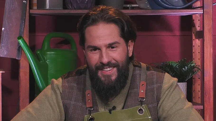 Maurice Dziwak hat „Promi Big Brother“ gewonnen Maurice Dziwak lächelt mit Bart und grüner Schürze mit der Aufschrift „Gartenleitung“. Hinter ihm stehen Gießkannen im Regal.
