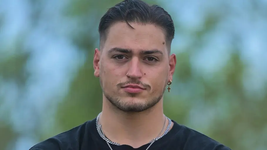 Max Bornmann Max Bornmann trägt ein schwarzes T-Shirt, silberne Ketten, Nasenring und Kreuzohrring vor unscharfem grün-blauem Hintergrund.
