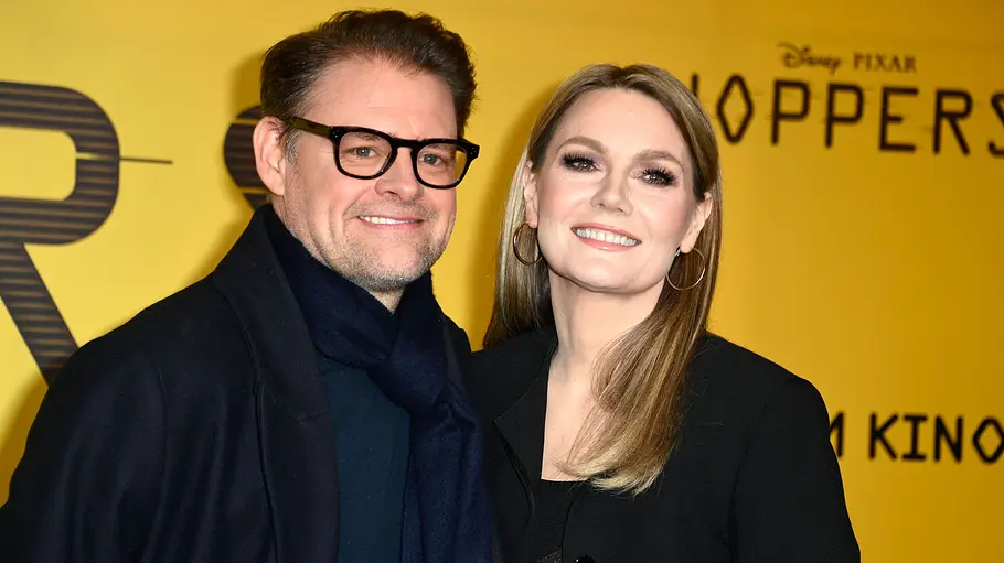 Max Giermann und Martina Hill Max Giermann mit Brille Marina Hill mit langen blonden Haaren stehen vor einem gelben Hintergrund, beide lächeln in die Kamera.
