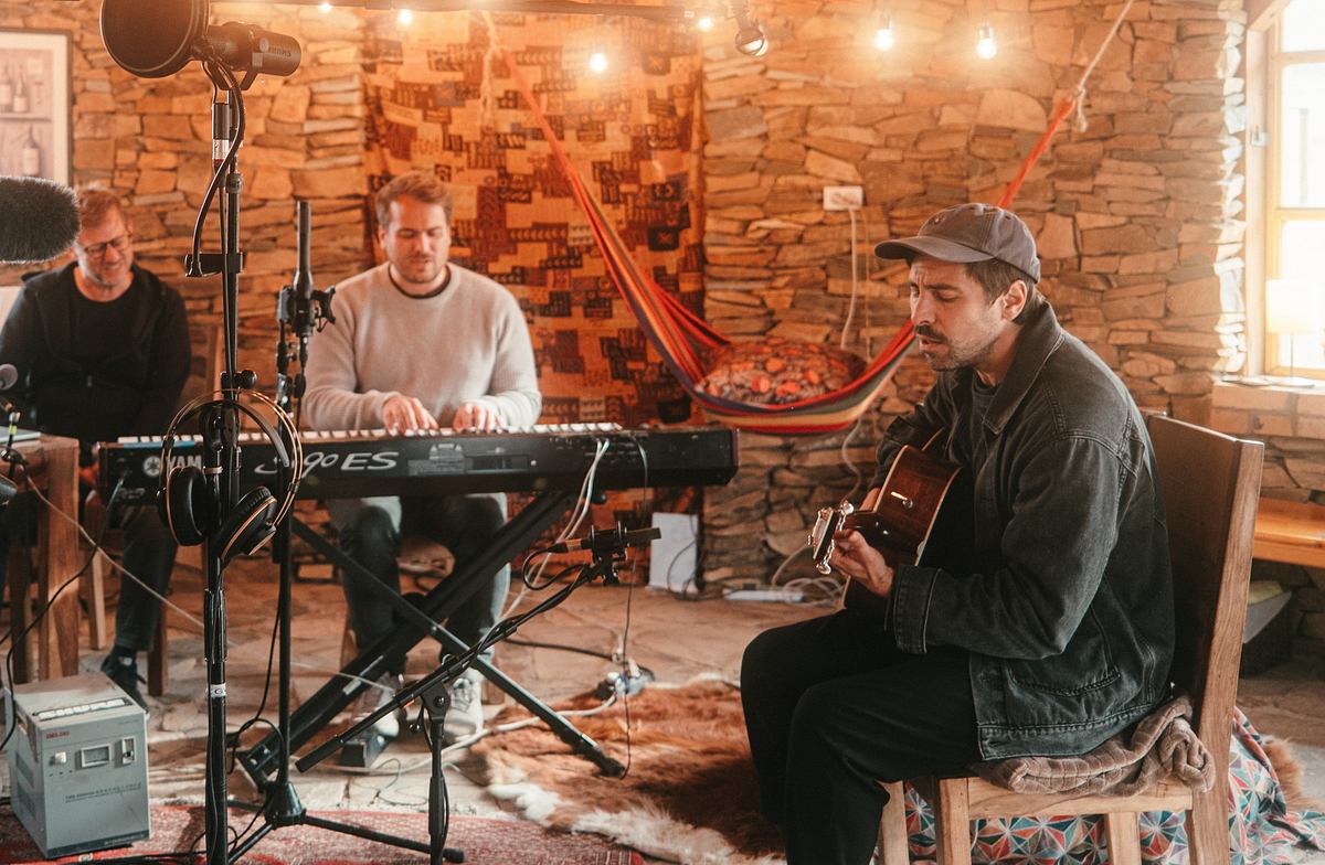 Song Trip: Max Giesinger x Mongolei: Musiker Max Giesinger sitzt im Studio und spielt Gitarre. Zwei weitere Männer sitzen im Studio, einer spielt Keyboard. Der Raum ist orange beleuchtet. Im Hintergrund hängt eine bunte Hängematte.