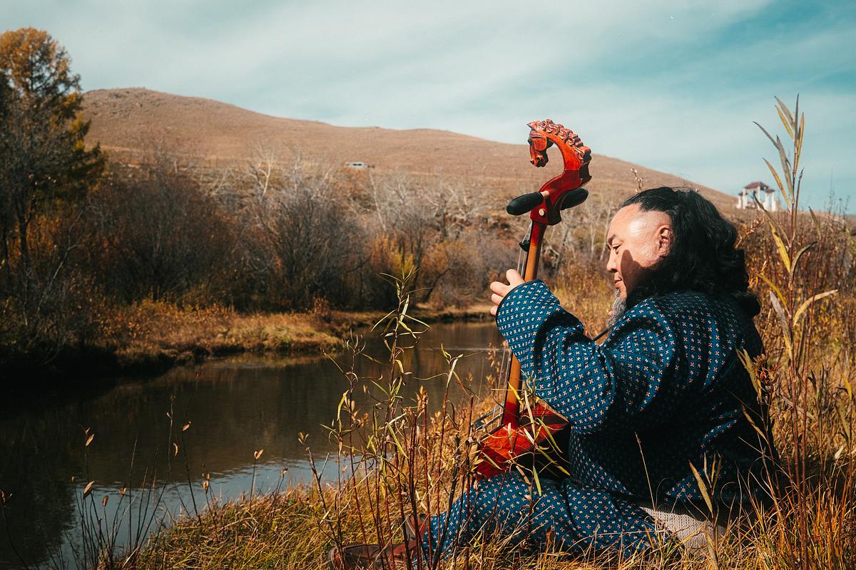 Song Trip: Max Giesinger x Mongolei: Der mongolische Musiker und Kehlkopfsänger Batzorig Vaanchig sitzt mit seinem Instrument auf einer Wiese am Ufer eines Gewässers. Er spielt die Pferdekopfgeige. Er trägt ein blaues Gewand und hat schwarze lange Haare.