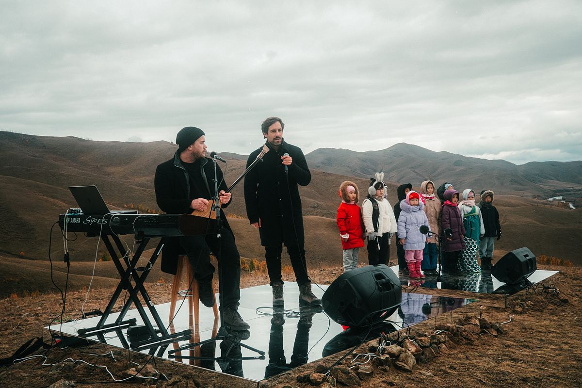 Song Trip: Max Giesinger x Mongolei: In einer kargen weiten Hügellandschaft: Ein Mann sitzt vor einem Keyboard auf einem Stuhl. In seiner Hand hält er ein Instrument. Neben ihm steht Max Giesinger und hält ein Mikrofon. Daneben stehen neun Kinder eines Kinderchors.