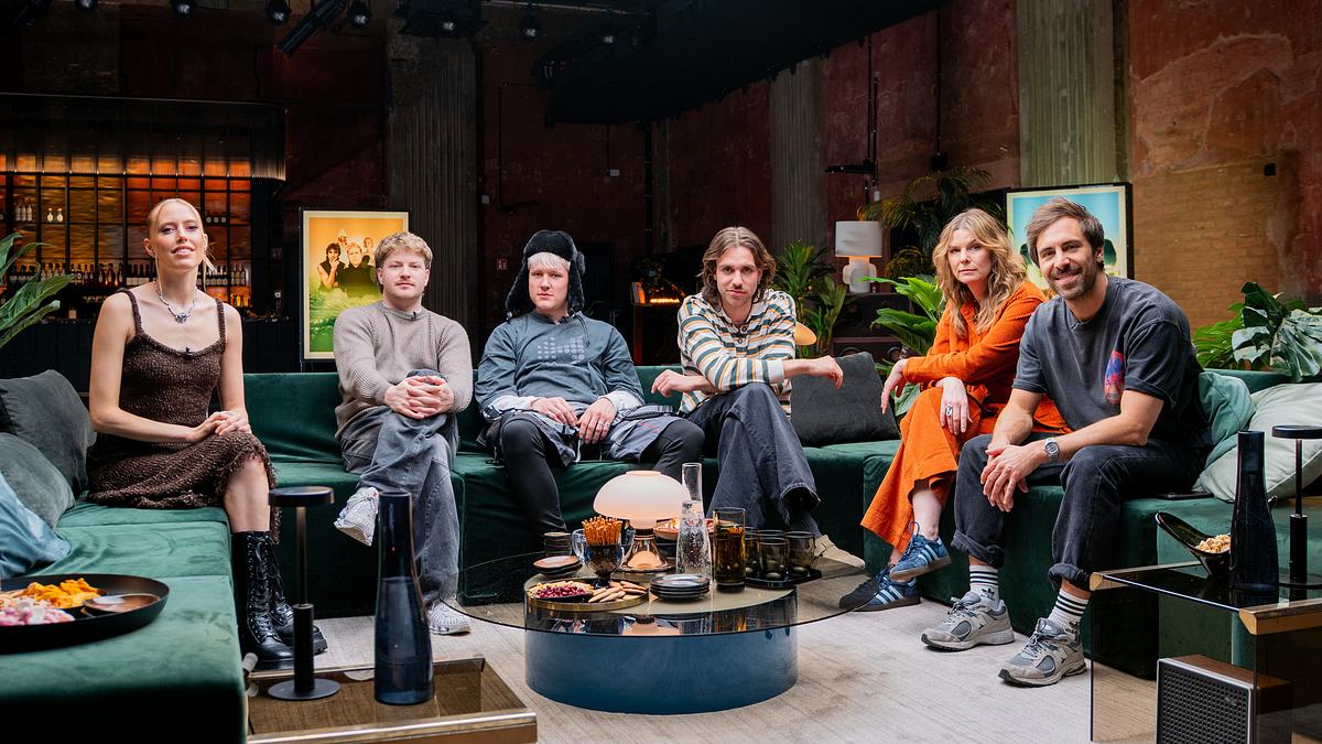 Der Cast von Song Trip von links: LEA, 01099, Eva Briegel von der Band Juli und Max Giesinger Song Trip: Max Giesinger x Mongolei: Sechs Personen sitzen auf einem grünen Ecksofa in einem modernen, schwach beleuchteten Raum mit freiliegenden Backsteinwänden: LEA, drei Mitglieder der Band 01099, Eva Briegel von der Band Juli und Max Giesinger. Vor ihnen steht ein runder Couchtisch mit verschiedenen Gegenständen, darunter Snacks, Getränke und Dekorationsobjekte. Im Hintergrund sind Pflanzen und gerahmte Kunstwerke zu sehen.