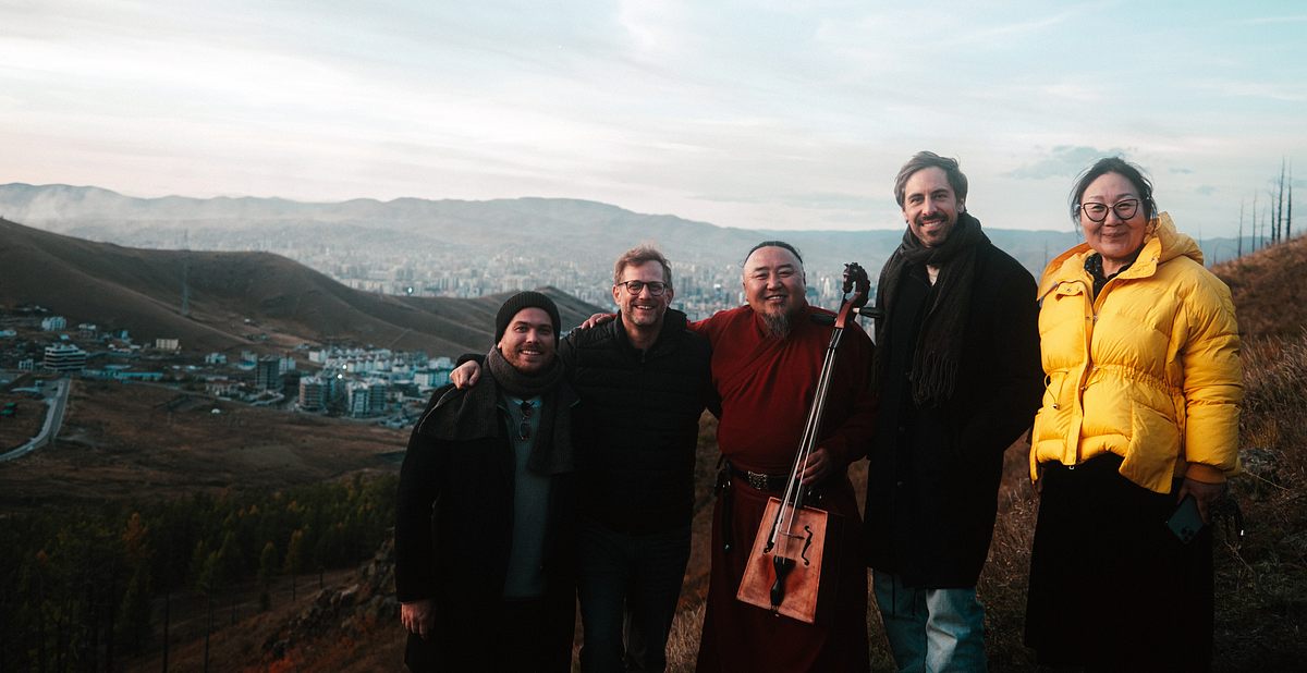 Song Trip: Max Giesinger x Mongolei: In einer weiten Berglandschaft mit einer Stadt im Hintergrund steht das Team von Max Giesinger mit ihm, dem Musiker Batzorig Vaanchig und der einheimischen Reiseführerin Chimeg auf einer Anhöhe. Alle schauen lächelnd in die Kamera.