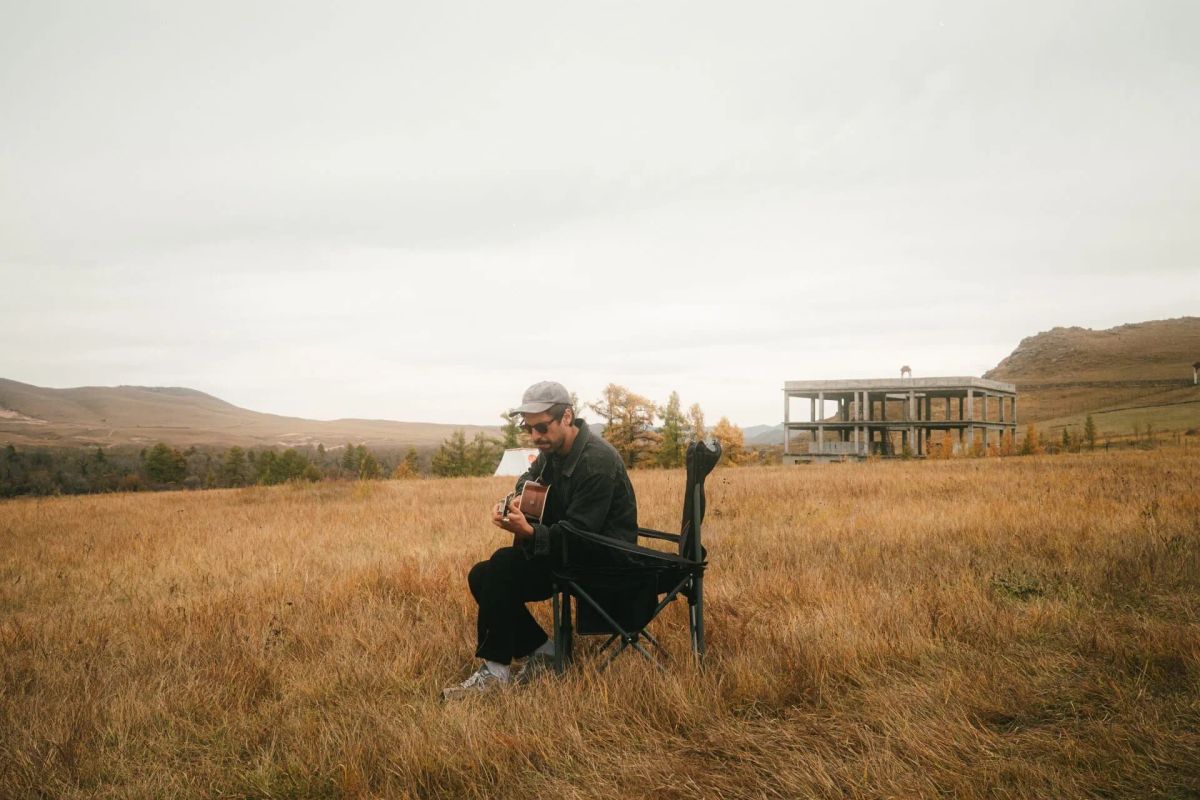 Song Trip: Max Giesinger x Mongolei: Musiker Max Giesinger sitzt mitten auf einem Feld auf einem Klappstuhl und spielt Gitarre.