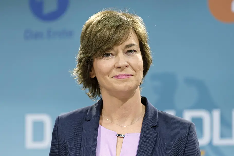 Maybrit Illner Maybrit Illner mit kurzen braunen Haaren trägt einen dunkelblauen Blazer über einer fliederfarbenen Bluse vor blauem Hintergrund.