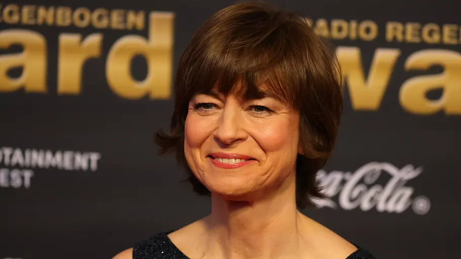Maybrit Illner und ihre Ex-Männer. Maybrit Illner mit kurzen braunen Haaren trägt ein dunkles, glitzerndes Oberteil vor einer Wand mit Schriftzügen.