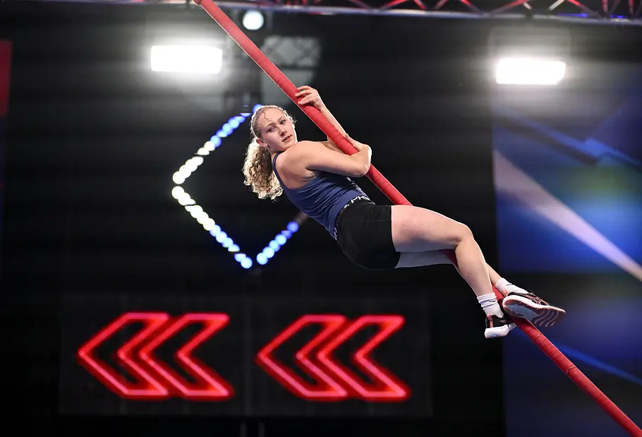 Megan Schüttler ist Teilnehmer bei Ninja Warrior Germany 2026 Megan Schüttler hält sich mit Armen und Beinen an einer schrägen roten Stange fest. Dahinter leuchten rote Pfeile.