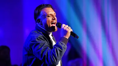 Menowin performt mit geschlossenen Augen und Mikrofon in der Hand auf einer Bühne mit blau-violetter Beleuchtung. - Foto: RTL / Stefan Gregorowius