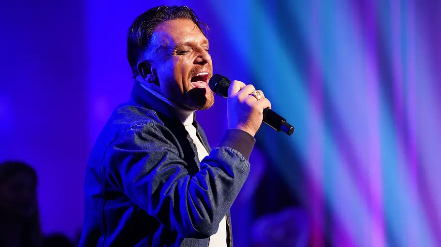 Menowin Fröhlich Menowin performt mit geschlossenen Augen und Mikrofon in der Hand auf einer Bühne mit blau-violetter Beleuchtung.