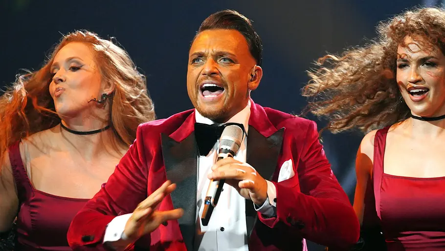 Menowin Fröhlich in der zweiten Live-Show Menowin im roten Samt-Sakko mit Fliege steht auf einer Bühne und hält ein Mikrofon, links und rechts von ihm tanzen zwei Frauen.