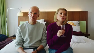 Merz gegen Merz: Geständnisse: Erik (Christoph Maria Herbst) sitzt neben Anne (Annette Frier) auf einem Hotelbett. Auf seinem Schoß liegt ein Notizblock und er schaut konzentriert nach vorne. Sie hält ein Sektglas und blickt zur Seite. - Foto: ZDF/Brendan Uffelmann