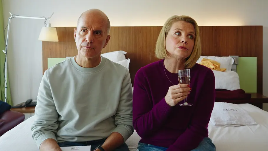 Anne Merz (Annette Frier) und ihr Ex-Mann Erik (Christoph Maria Herbst) nutzen den kurzen Moment der Ruhe, bevor das Chaos der anderen Familienmitglieder wieder losbricht. Merz gegen Merz: Geständnisse: Erik (Christoph Maria Herbst) sitzt neben Anne (Annette Frier) auf einem Hotelbett. Auf seinem Schoß liegt ein Notizblock und er schaut konzentriert nach vorne. Sie hält ein Sektglas und blickt zur Seite.