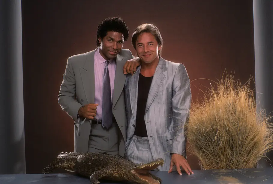 Miami Vice Don Johnson und Philip Michael Thomas als Crockett und Tubbs in „Miami Vice“, sie stehen nebeneinander im Studio und posieren vor einem Tisch mit einem Alligator