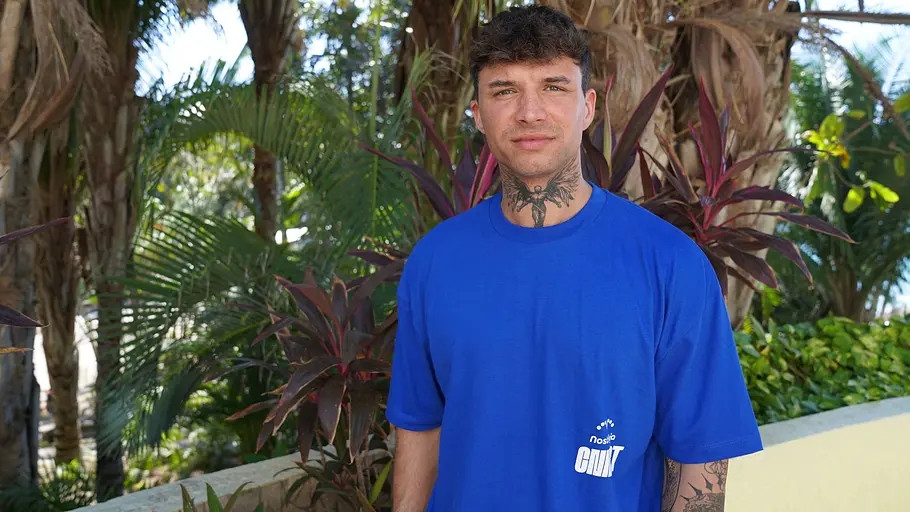 Micha steht in einem blauen T-Shirt vor tropischer Kulisse. 