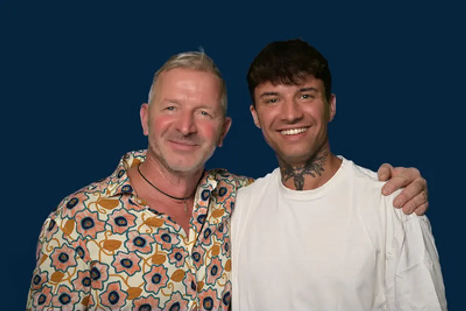 Micha im weißen T-Shirt und sein Vater Micha Klotz im bunten Hemd stehen lächelnd Arm in Arm vor dunkelblauem Hintergrund.