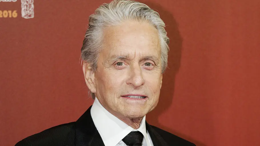Michael Douglas Affären Michael Douglas mit grauem Haar trägt einen schwarzen Anzug, weißes Hemd und schwarze Krawatte vor rotem Hintergrund.