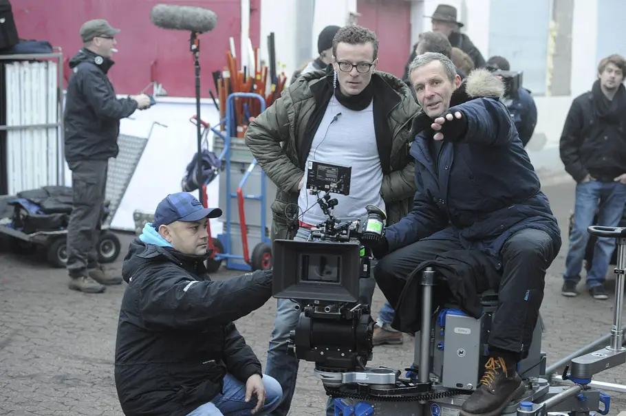 Michael Illner Drehbuchautor Drei Männer arbeiten an einer Filmkamera auf einem Kamerawagen. Einer zeigt nach vorn, im Hintergrund steht weitere Filmcrew.