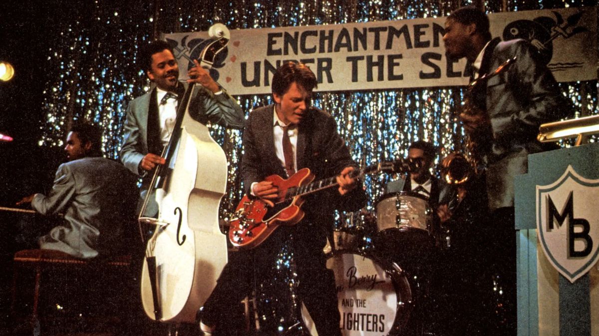 Zurück in die Zukunft Man sieht Michael J. Fox in einem Anzug auf einer Bühne in einer High School Aula. Er trägt eine rötliche Krawatte und hinter ihm ist eine Band, bestehend aus vier dunkelhäutigen Mitgliedern, die ein Piano, Kontrabass, Schlagzeug und einen Bass spielen. Ihre Anzüge sind universell und gehen ins dunkle Blau über.