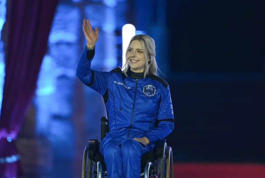 Michaela Benthaus Michaela Benthaus sitzt im Rollstuhl und hebt vor blau beleuchtetem Hintergrund eine Hand zum Gruß.