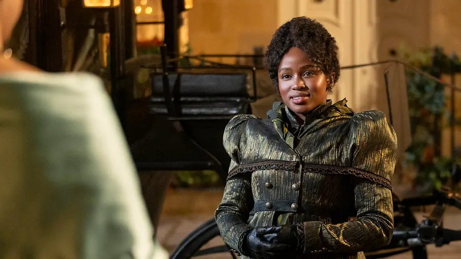 Michaela (Masali Baduza) in „Bridgerton“ Eine Frau mit dunkler Haut und lockigen Haaren steht in einem prunkvollen, historisch anmutenden Kleid vor einer Kutsche. Sie trägt schwarze Handschuhe und schaut lächelnd eine andere Person an, die nur teilweise im Bild zu sehen ist. Die Szene spielt bei Nacht, vor einem hell erleuchteten Gebäude.