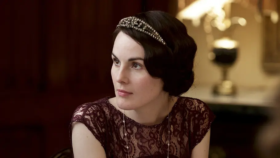 Michelle Dockery als Lady Mary Crawley Michelle Dockery in historischer Kleidung in einer Szene aus der Serie „Downton Abbey“, sitzend an einem Tisch in vornehmem Ambiente.