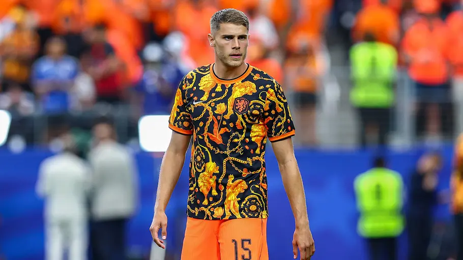 Fußballspieler der niederländischen Nationalmannschaft im schwarz-orange gemusterten Aufwärmtrikot und orangefarbenen Shorts läuft vor einem Spiel über den Rasen. Im Hintergrund sind Fans und das Stadion unscharf zu erkennen.