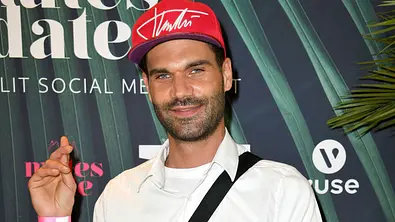 Mike Cees posiert bei einem Event vor einer Sponsorenwand, trägt ein weißes Hemd, eine rote Cap und eine Umhängetasche. - Foto: IMAGO / Future Image