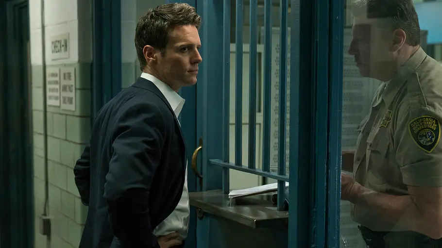 Mindhunter Ein Mann im Anzug spricht an einem vergitterten Schalter mit einem Justizbeamten in einem Gefängnis. Die Szene stammt aus der Krimiserie „Mindhunter“.