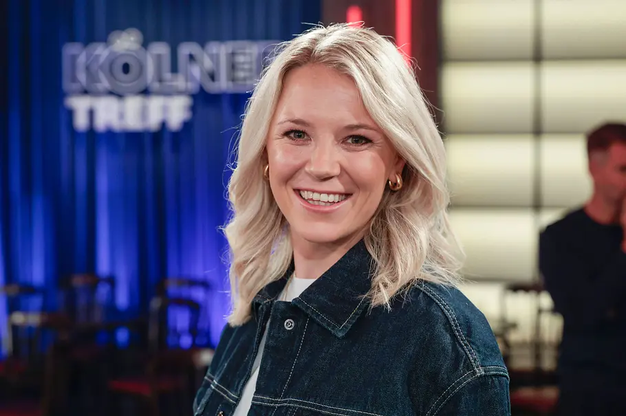 Miriam Neureuther Miriam Neureuther lächelt in einer dunklen Jeansjacke vor einem blauen Vorhang mit dem Schriftzug „Kölner Treff“.
