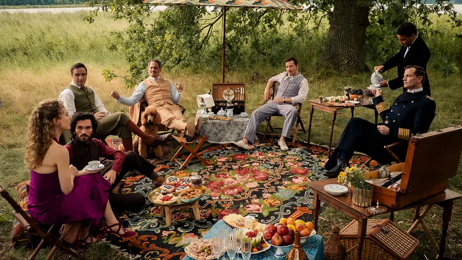 Eine Gruppe elegant gekleideter Menschen sitzt bei einem Picknick auf einer bunten Decke im Grünen unter Sonnenschirmen. Auf den Tischen und vor ihnen stehen Teller mit Obst, Gebäck und Getränken. Ein Hund sitzt bei einem der Männer, ein Butler schenkt Tee ein. Im Hintergrund ist eine Wiese mit Bäumen und ein See zu sehen.