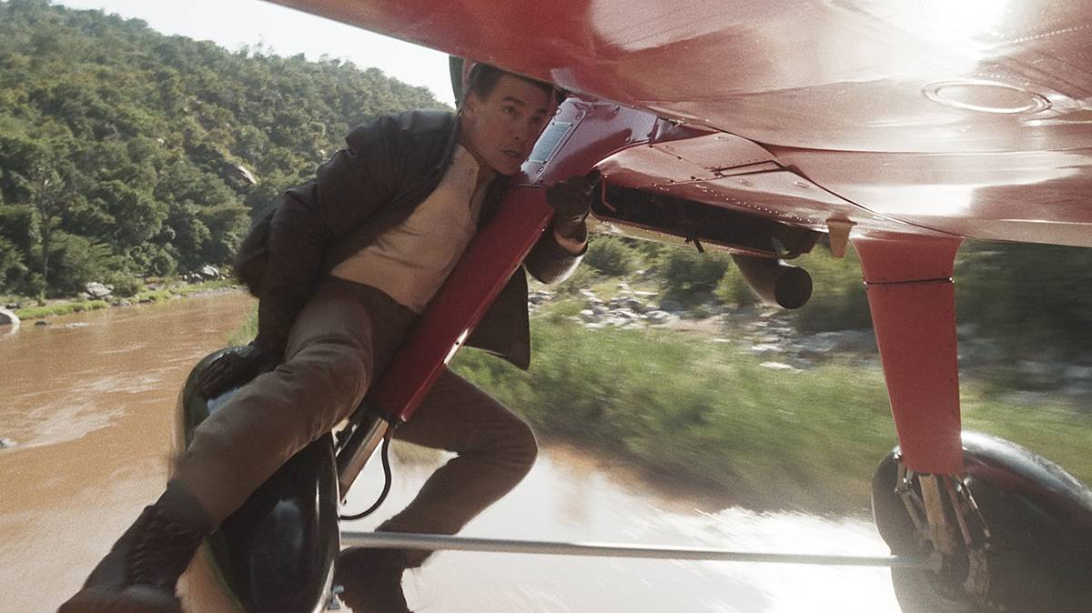 Für Piloten ist es vor dem Abflug ratsam, die Räder zu kontrollieren - Tom Cruise hängt vermutlich an ihnen. Tom Cruise klammert sich an ein fliegendes, rotes Propellerflugzeug. Er blickt angespannt nach vorne.