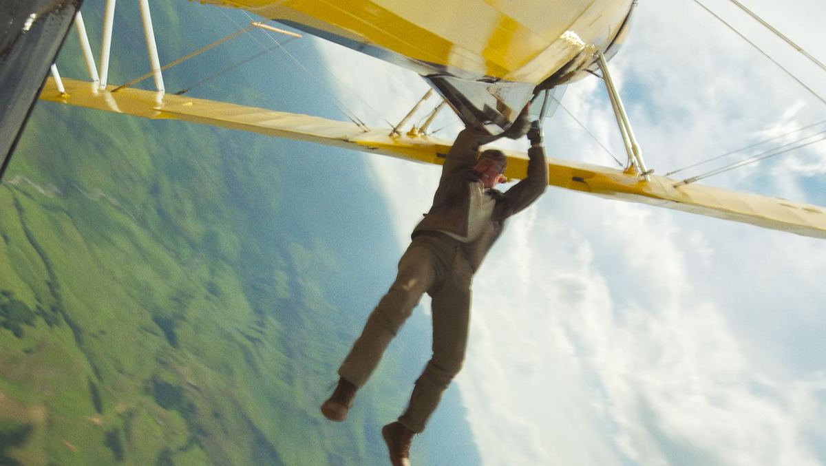 Auch wenn die Bilder in diesem Artikel einen anderen Eindruck vermitteln: Tom Cruise fliegt auch gerne sitzend in Flugzeugen. Tom Cruise klammert sich an ein kopfüber fliegendes, gelbes Propellerflugzeug.