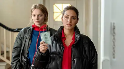 Mila und Anna Nowak stehen im Treppenhaus. Letztere trägt eine schwarze Steppjacke und zeigt einen Polizeiausweis. - Foto: © NDR/ARD/Thorsten Jander Mila und Anna Nowak stehen im Treppenhaus. Letztere trägt eine schwarze Steppjacke und zeigt einen Polizeiausweis. - Foto: © NDR/ARD/Thorsten Jander