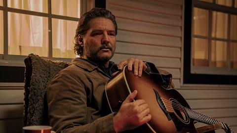 Pedro Pascal als Joel. Er sitzt mit einer Gitarre auf dem Schoß auf der Veranda. - Foto: Liane Hentscher/HBO