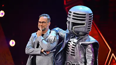 Matthias Opdenhövel steht neben Mr. Mic im silbernen Mikrofonkostüm, der einen Arm auf seine Schulter legt. - Foto: Joyn Matthias Opdenhövel steht neben Mr. Mic im silbernen Mikrofonkostüm, der einen Arm auf seine Schulter legt. - Foto: Joyn
