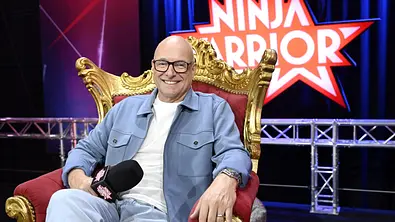 Ein lächelnder Mann mit Brille sitzt entspannt auf einem gold-roten Sessel und hält ein Mikrofon in der Hand. Im Hintergrund ist das „Ninja Warrior“-Logo auf einer Bühne zu sehen. - Foto: RTL / Markus Hertrich