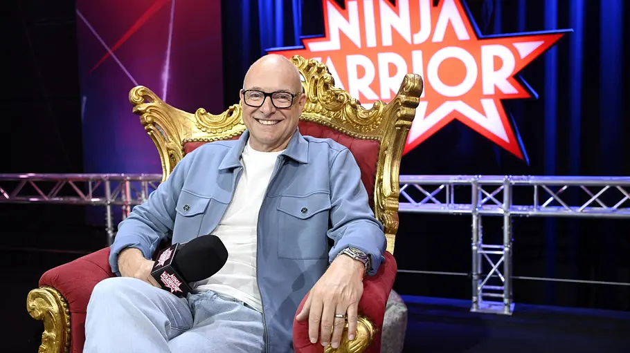 Moderator Frank Buschi Buschmann Ein lächelnder Mann mit Brille sitzt entspannt auf einem gold-roten Sessel und hält ein Mikrofon in der Hand. Im Hintergrund ist das „Ninja Warrior“-Logo auf einer Bühne zu sehen.