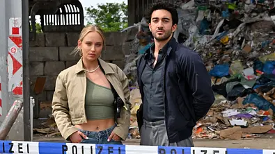 Szene aus „Mord im Revier“: Caro Cult als Lou und Eidin Jalali als Gianni stehen hinter Polizeiabsperrband vor einem Müllberg. - Foto: Foto: RTL / Hardy Spitz Szene aus „Mord im Revier“: Caro Cult als Lou und Eidin Jalali als Gianni stehen hinter Polizeiabsperrband vor einem Müllberg. - Foto: Foto: RTL / Hardy Spitz