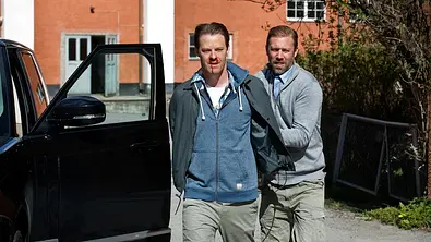 Ein Mann führt einen verletzten Verdächtigen neben einem schwarzen Auto ab. - Foto: © Johan Paulin/Filmlance International AB