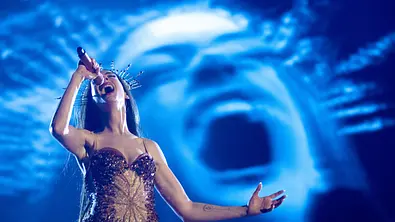 Anne Hathaway steht in einem spärlichen Kostüm auf einer Bühne und singt einen Song. Der Hintergrund ist blau und zeigt ihr Gesicht.  - Foto: Zoey Kang (A24)